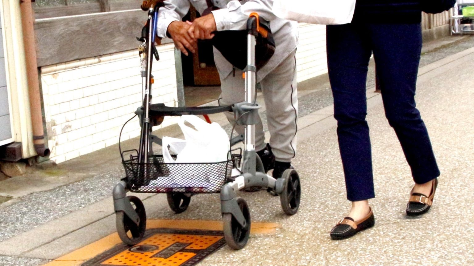 入居者が歩行器を使用しながら屋外で移動する様子。介護職員が付き添い支援している。