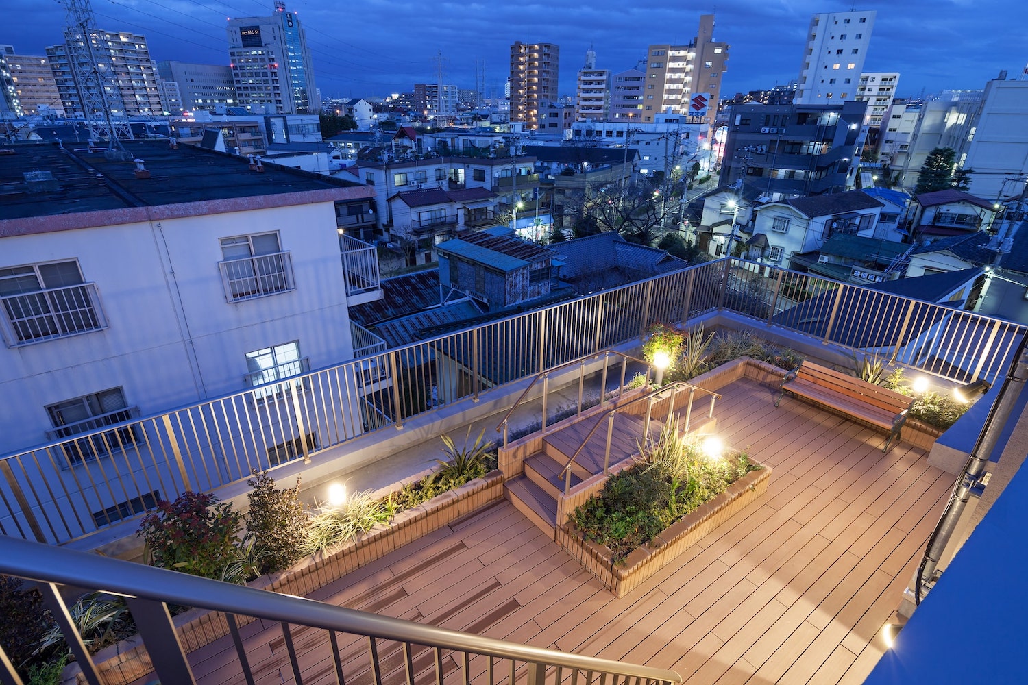 施設の屋上テラス。夜間照明で整備された木製デッキと植栽、都市景観が見える。